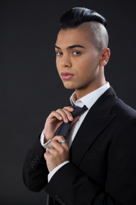 This masculine Latino person wears a black suit that fades into the black background. He adjusts his slim black tie and gazes casually into the camera.