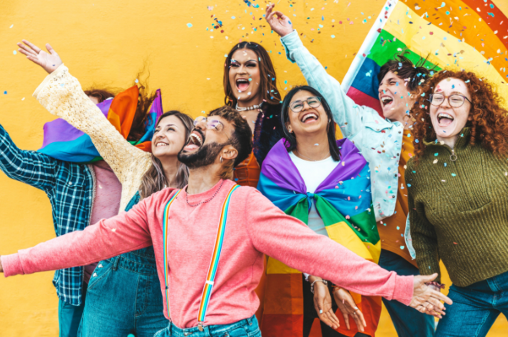 Seven people stand tightly together. Dressed in colorful clothing and waving pride flags or using pride flags as capes, the people radiate queer joy. They are smiling with open mouths and have outstretched arms.