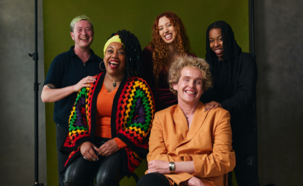 In this group photo, three people stand behind two seated people, all smiling or laughing. There are people of many gender expressions represented.